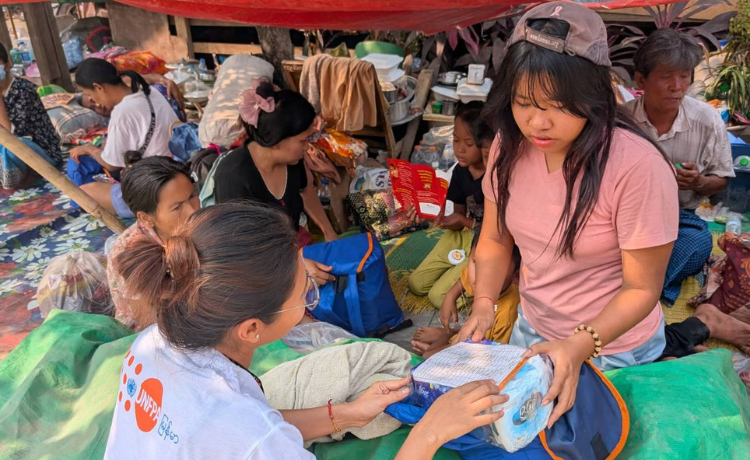 Dans un camp de fortune, une femme portant un t-shirt floqué de l’UNFPA tend un colis bleu à une jeune femme en t-shirt rose