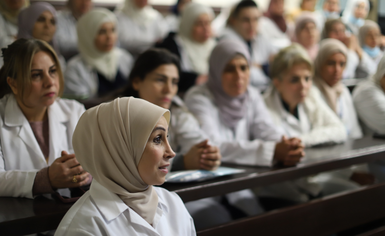  قابلة ترتدي معطفا أبيض وحجابا تتحدث في المقدمة، وخلفها العديد من القابلات الأخريات في المعاطف البيضاء.